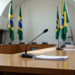 Mesa com microfone e bandeiras do Brasil em sala de assembleia legislativa